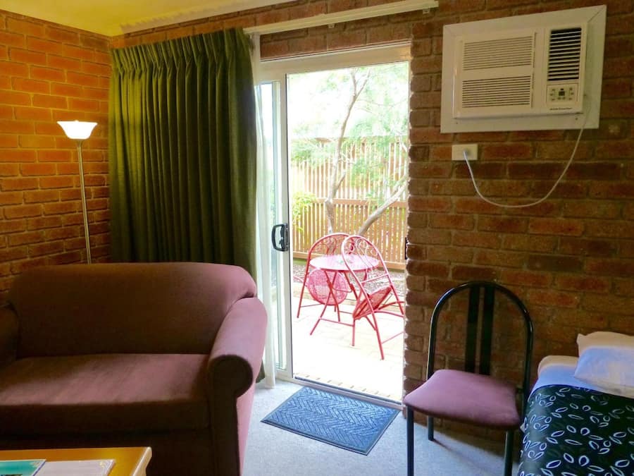 Une pièce avec un mur en briques, une porte-fenêtre coulissante donnant sur un espace extérieur avec une table et des chaises en métal rouge, un climatiseur blanc et un paillasson bleu.