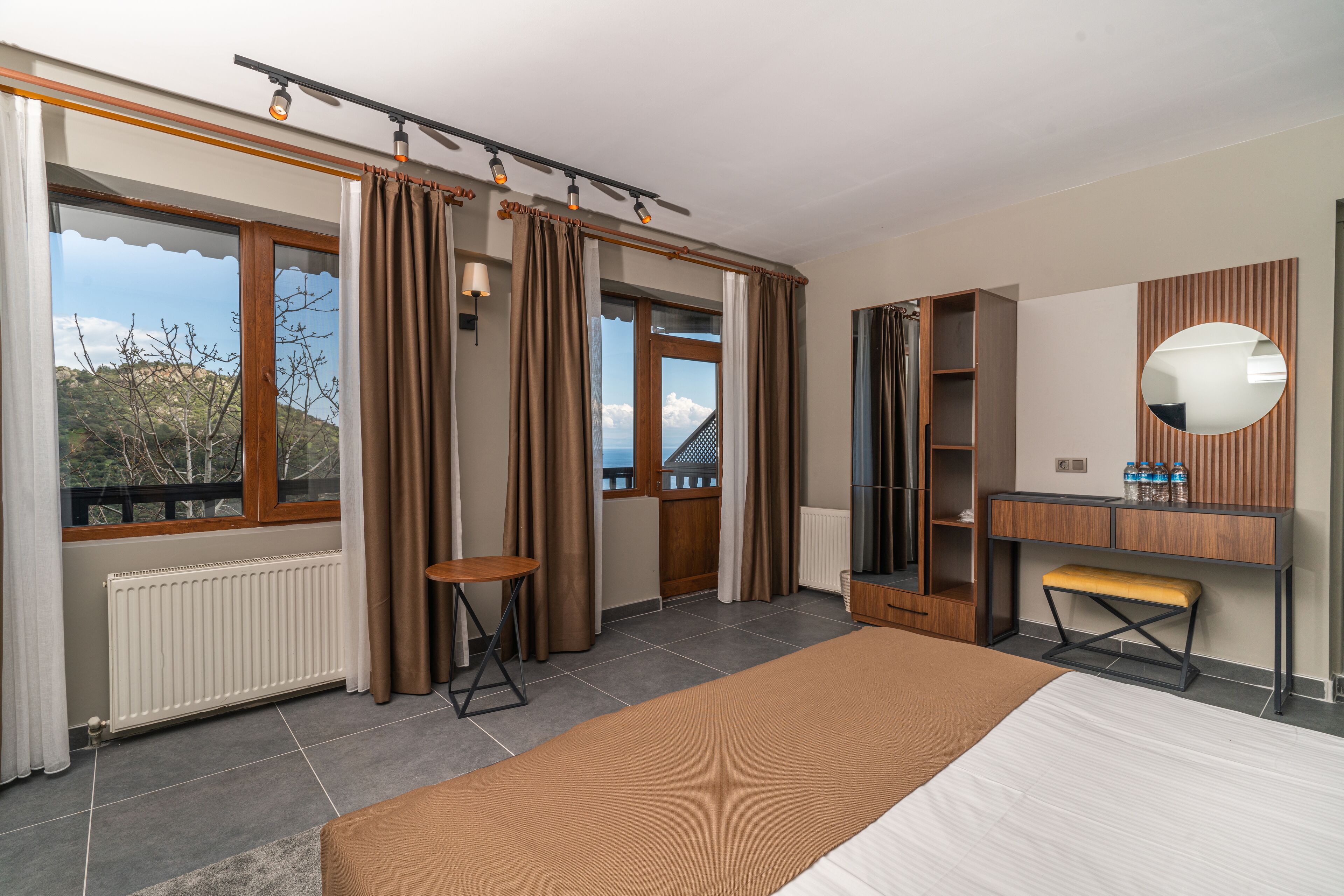 A modern bedroom with a large bed, a wooden bench, a mirror, and a view of the outdoors.
