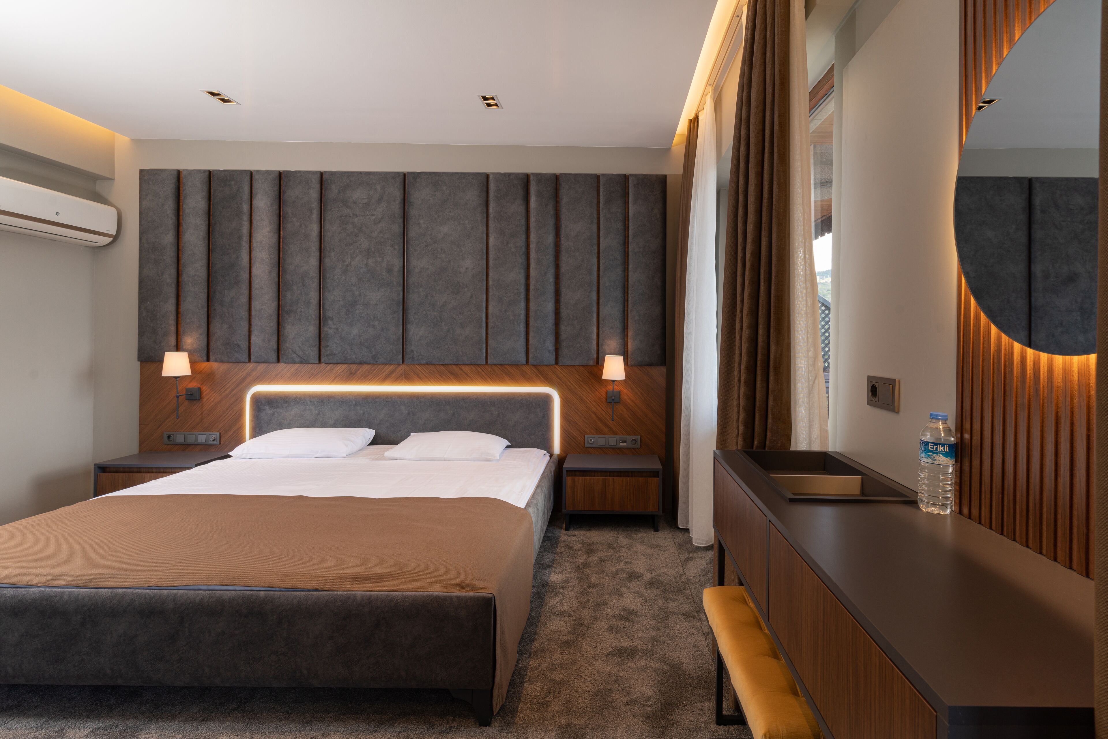 A modern hotel room with a large bed, bedside tables, a bench, a mirror, and a water bottle on the nightstand.