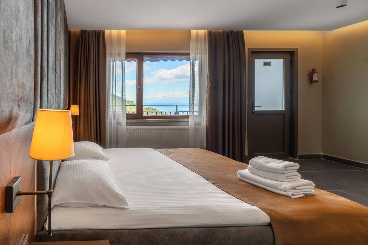 Une chambre d’hôtel avec un lit, une lampe, une fenêtre avec des rideaux, une porte et une pile de serviettes.