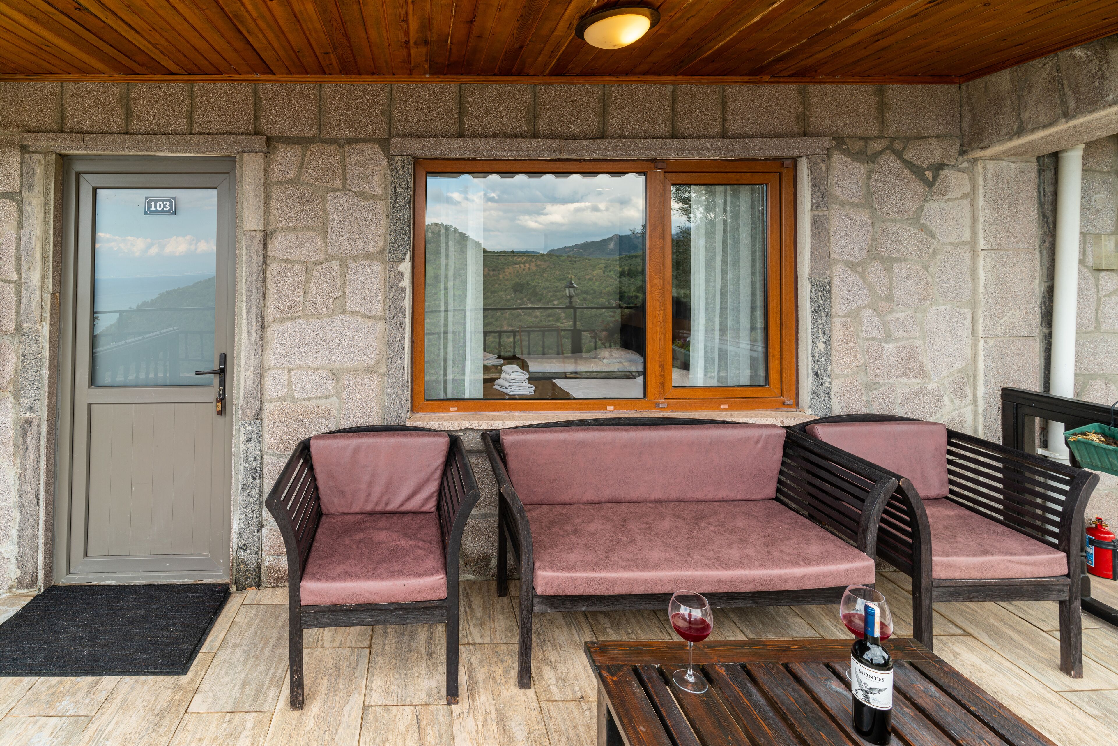 A stone-walled room with a wooden ceiling, a door marked with the number 103, a bench with pink cushions, and a table with a bottle of wine.