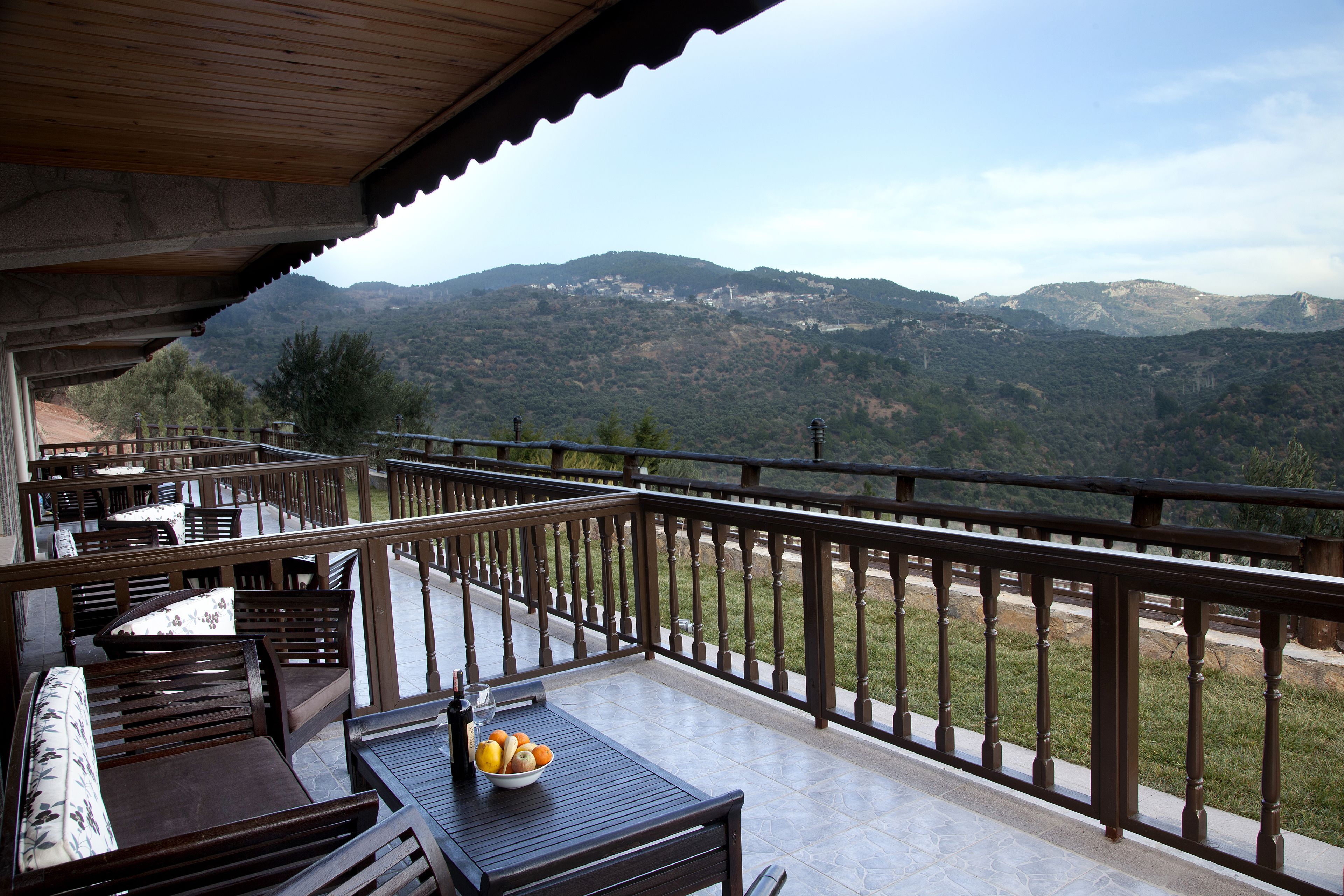 Un balcón con mobiliario de madera, una mesa con un frutero y una botella, y una vista panorámica de las montañas.