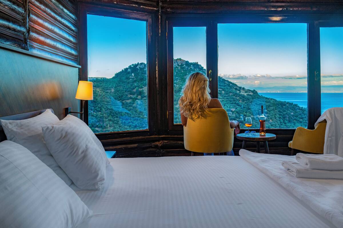 Une chambre d’hôtel avec un grand lit, une petite table sur laquelle se trouvent une bouteille et des verres, et une vue sur un paysage montagneux par de grandes fenêtres.