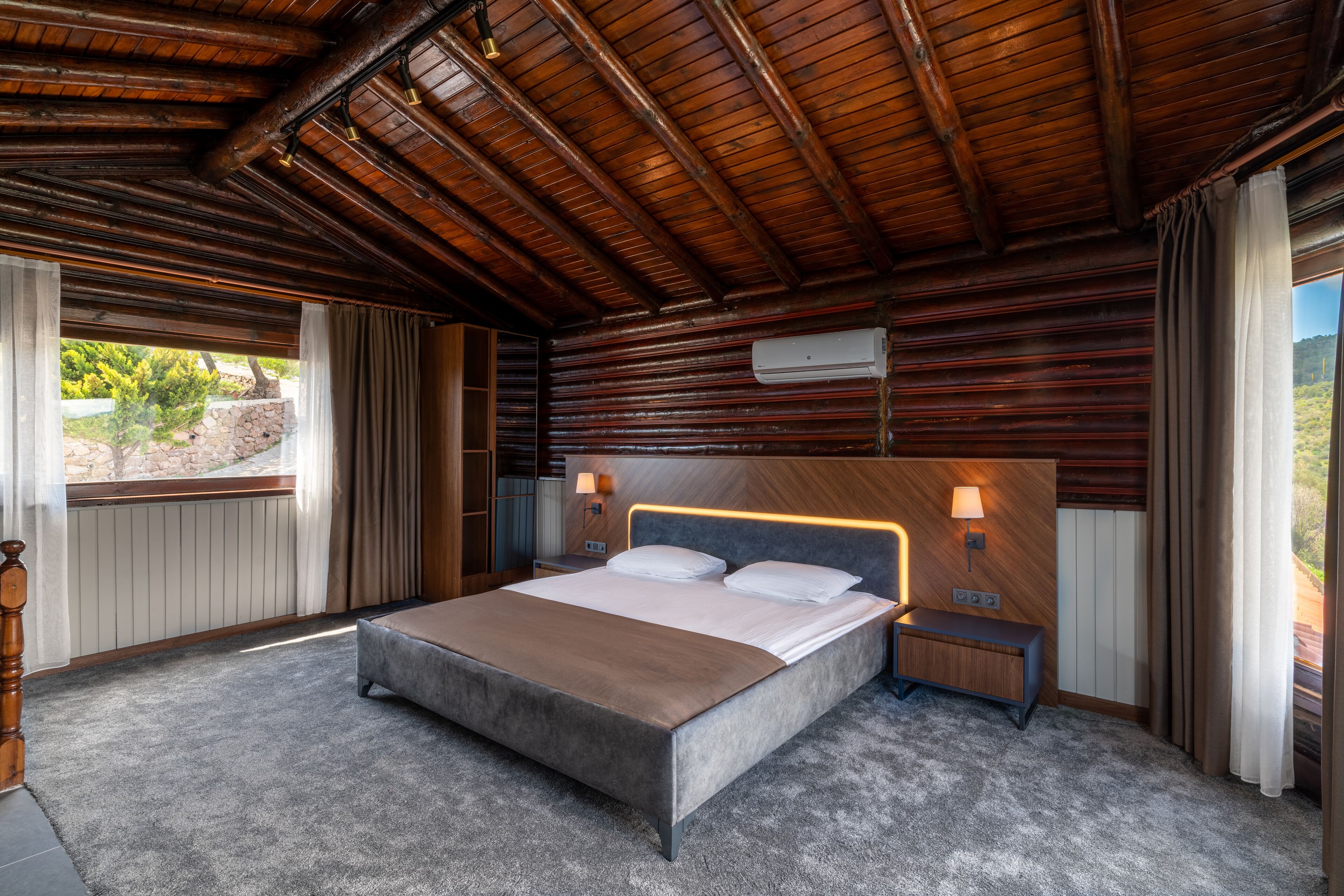 A bedroom with a large bed, wooden ceiling, and a view of greenery outside.