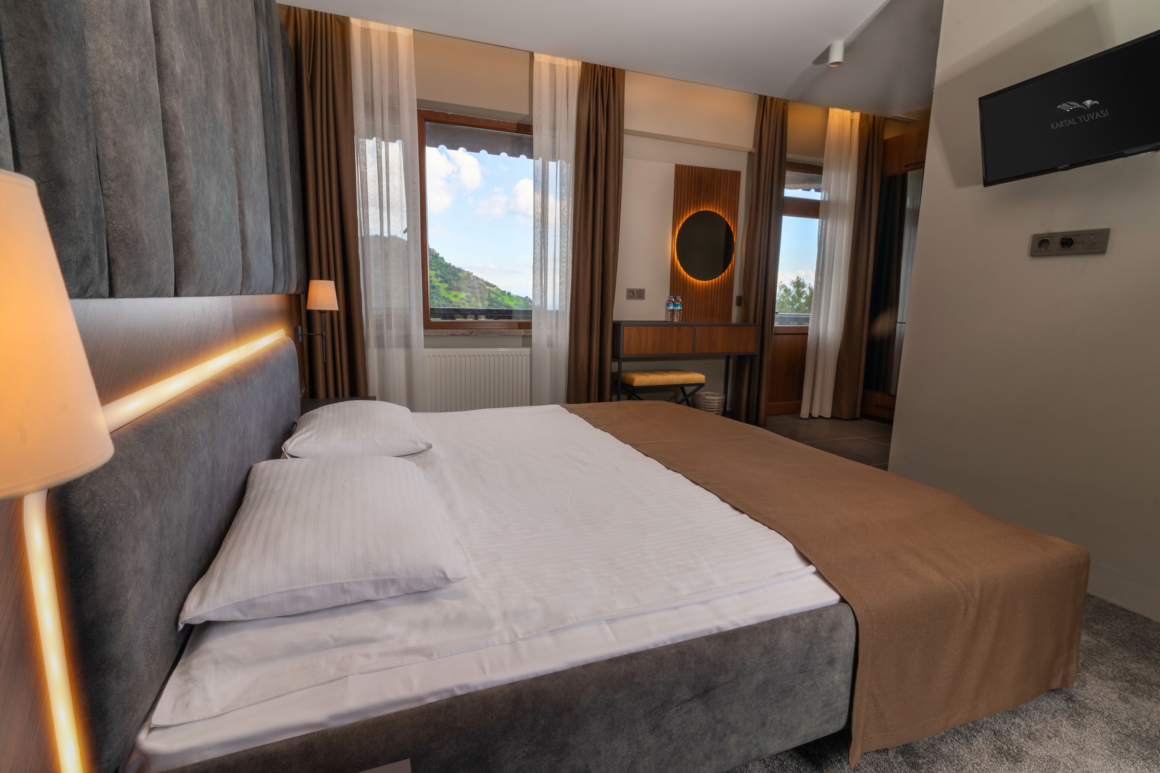 Quarto de hotel com uma cama grande, abajur, vista para a montanha, escrivaninha de madeira, espelho e televisão na parede.