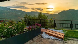 Une terrasse offrant une vue sur le coucher de soleil au-dessus des collines, un banc en bois et des parterres de fleurs.
