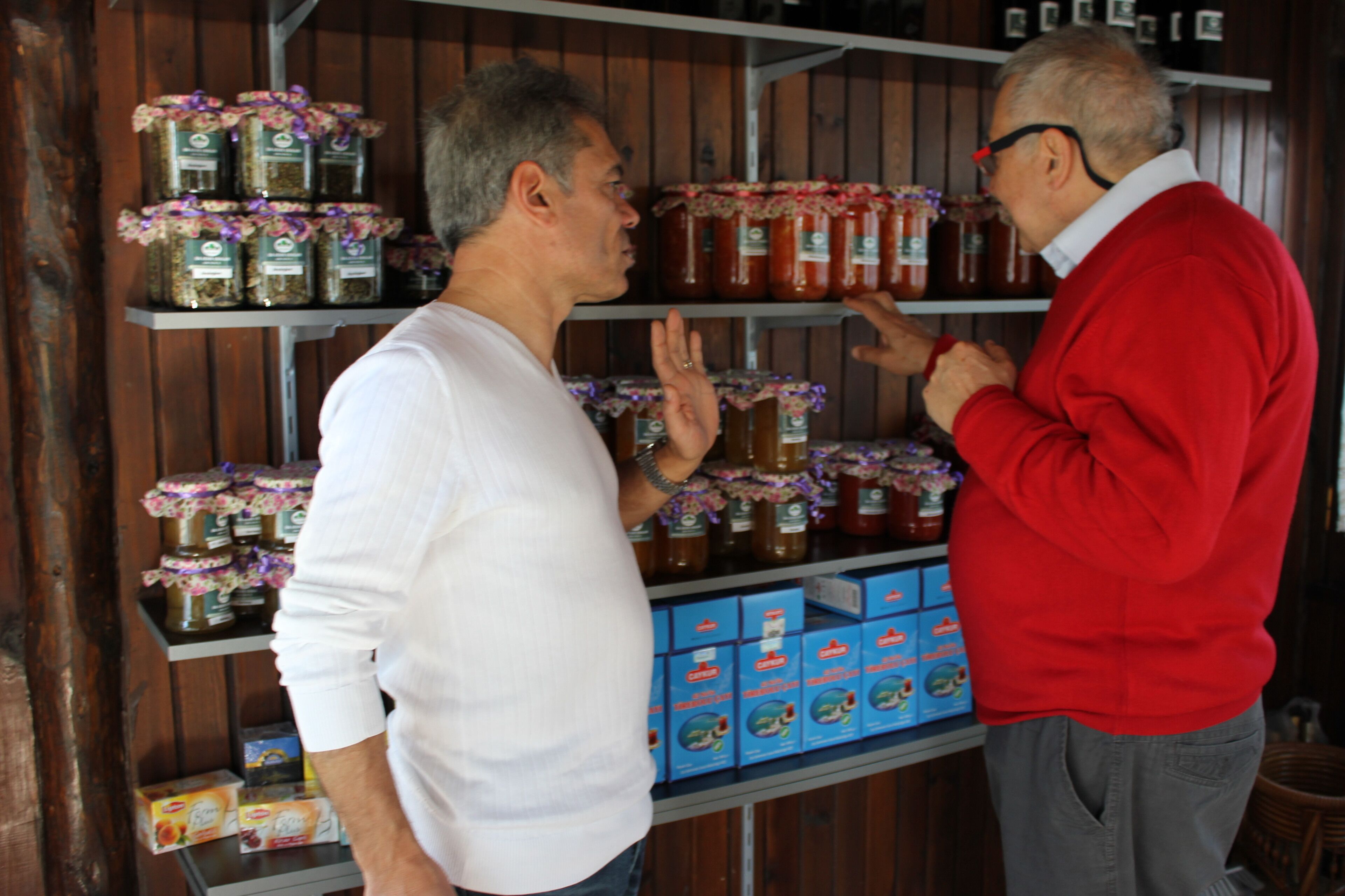 Two men in a rustic setting with shelves stocked with jars and boxes, likely a store or market.