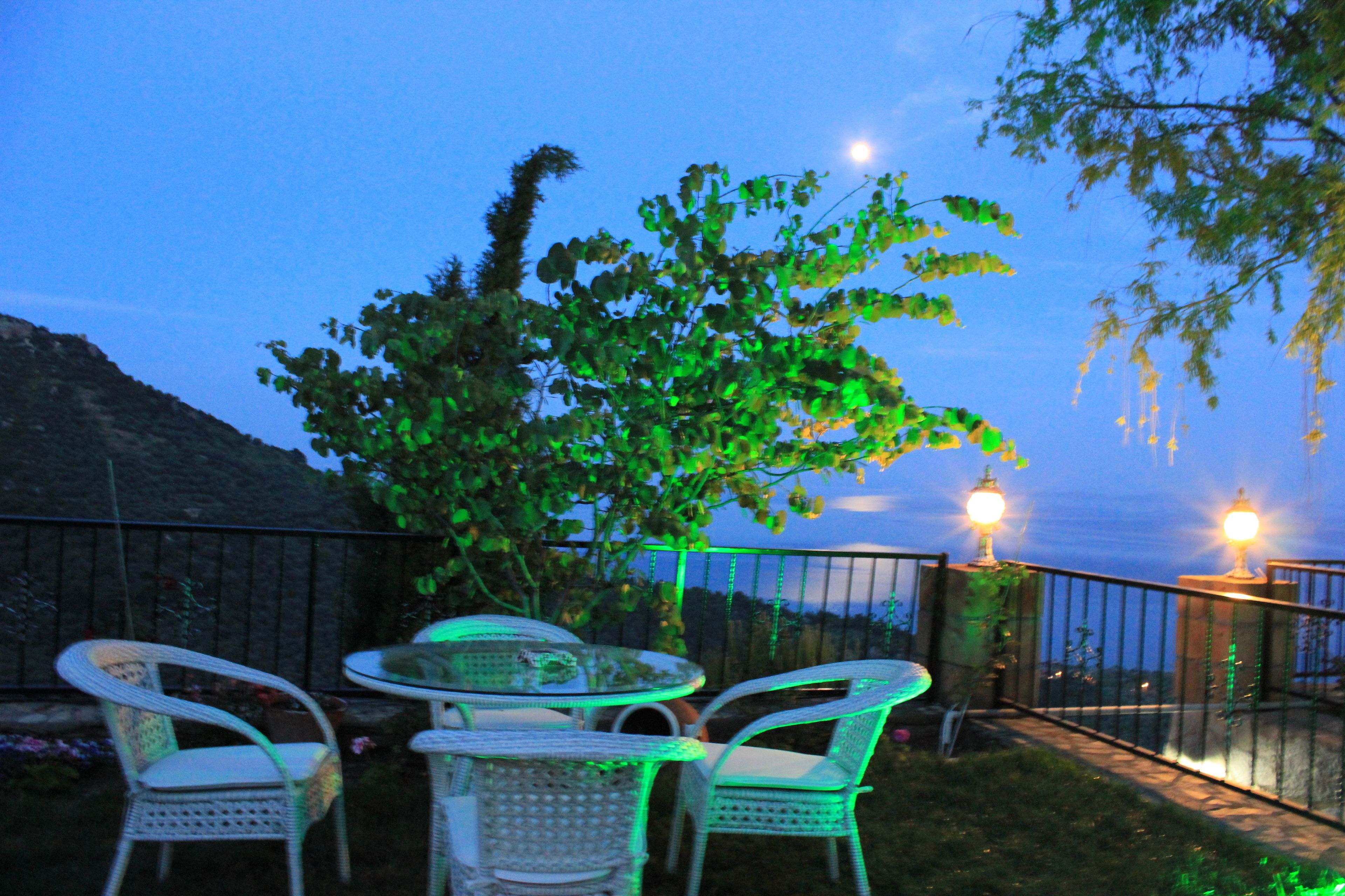 An outdoor patio with a round table and two chairs, a tree, and a railing with lights.