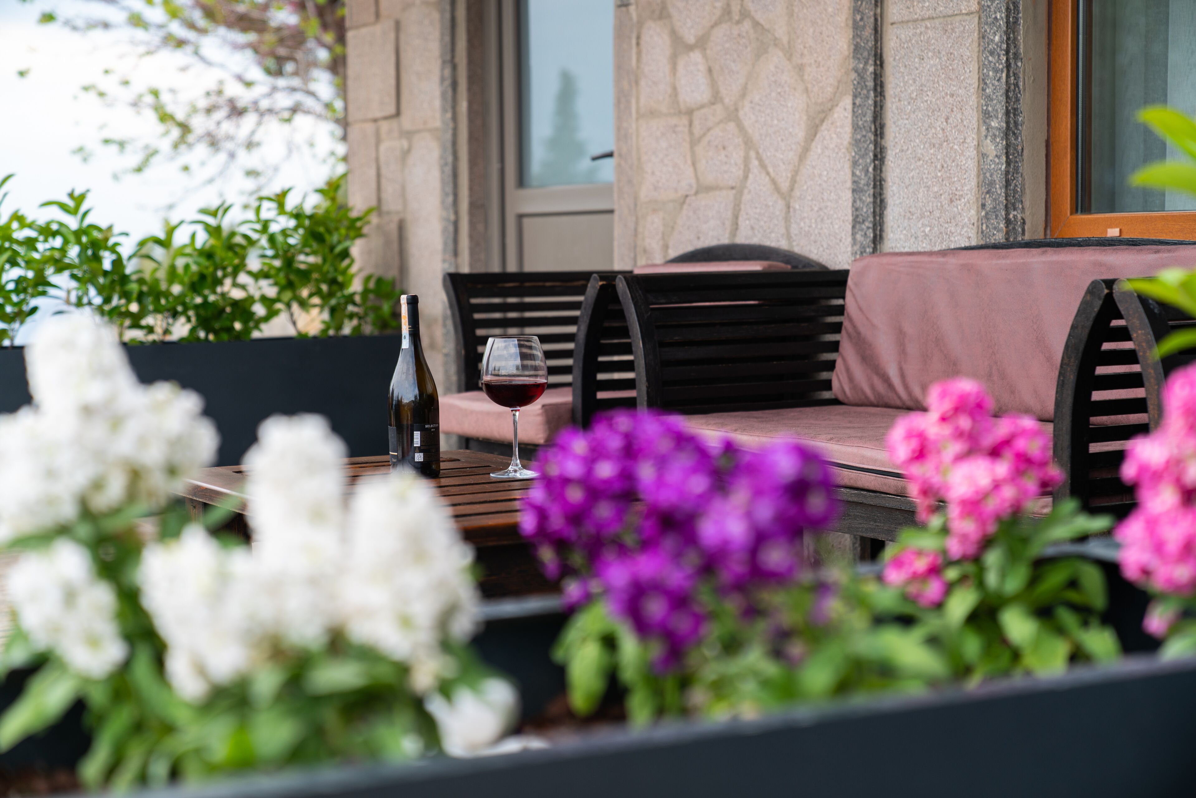 A patio with a wooden table, a wine bottle, and a glass of red wine. There are chairs and a cushioned bench.