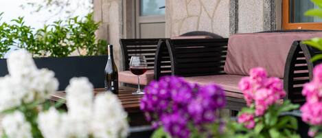 Un patio con una mesa de madera, una botella de vino y una copa de vino tinto. Hay sillas y un banco acolchado.