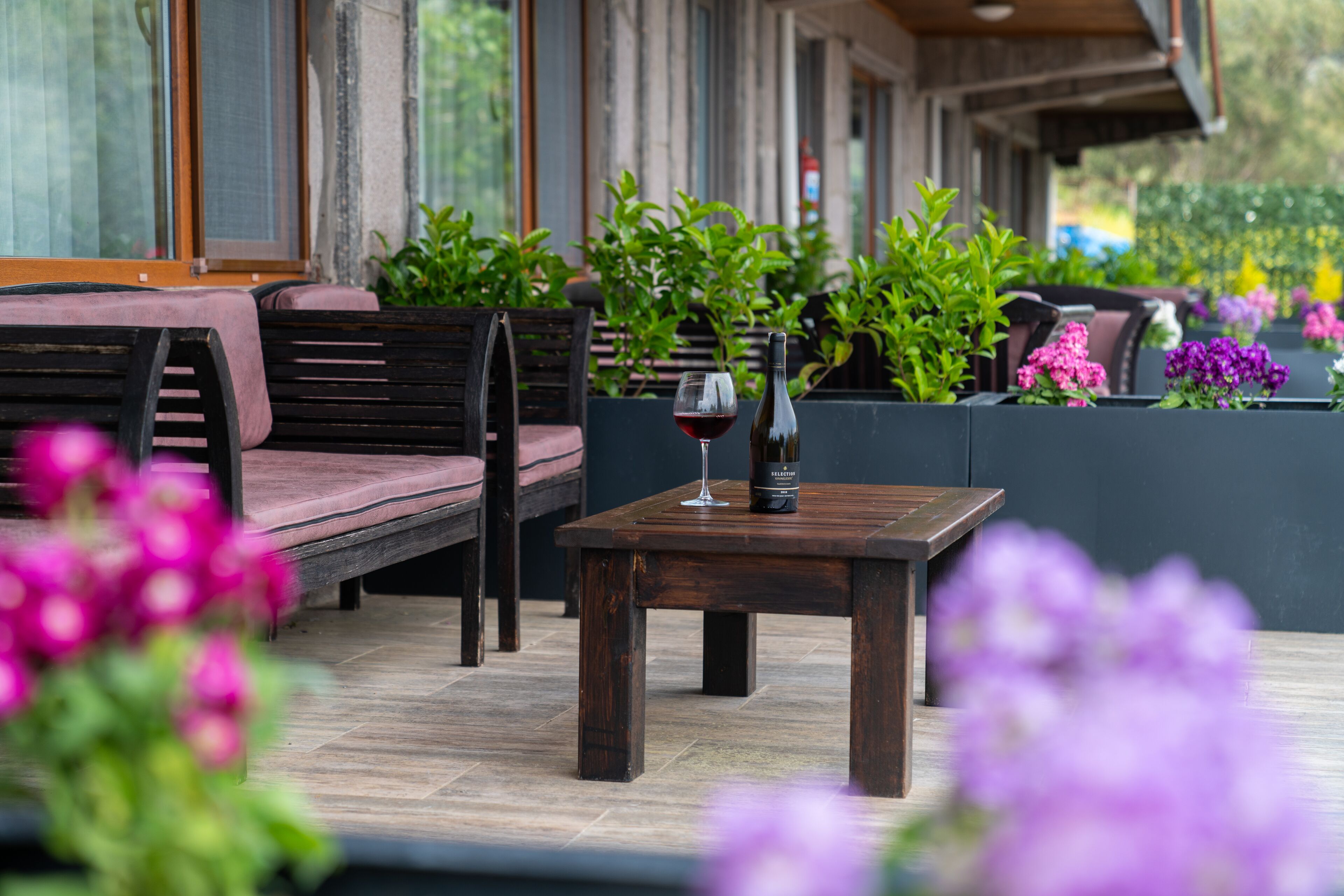 Ein Holztisch mit einer Weinflasche und einem Glas, umgeben von Grünpflanzen und violetten Blumen.