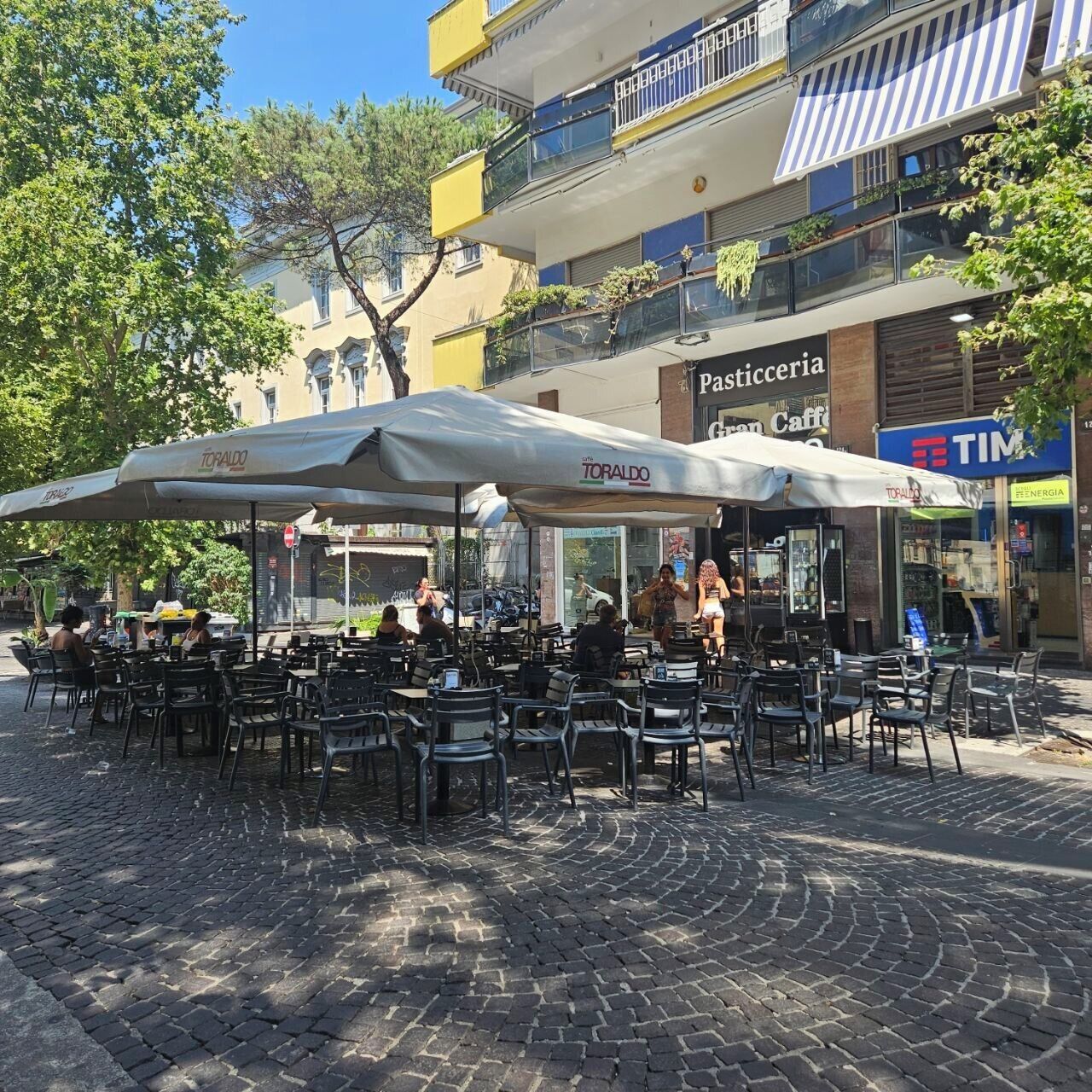 An outdoor café with tables and chairs, surrounded by buildings with shops and balconies.