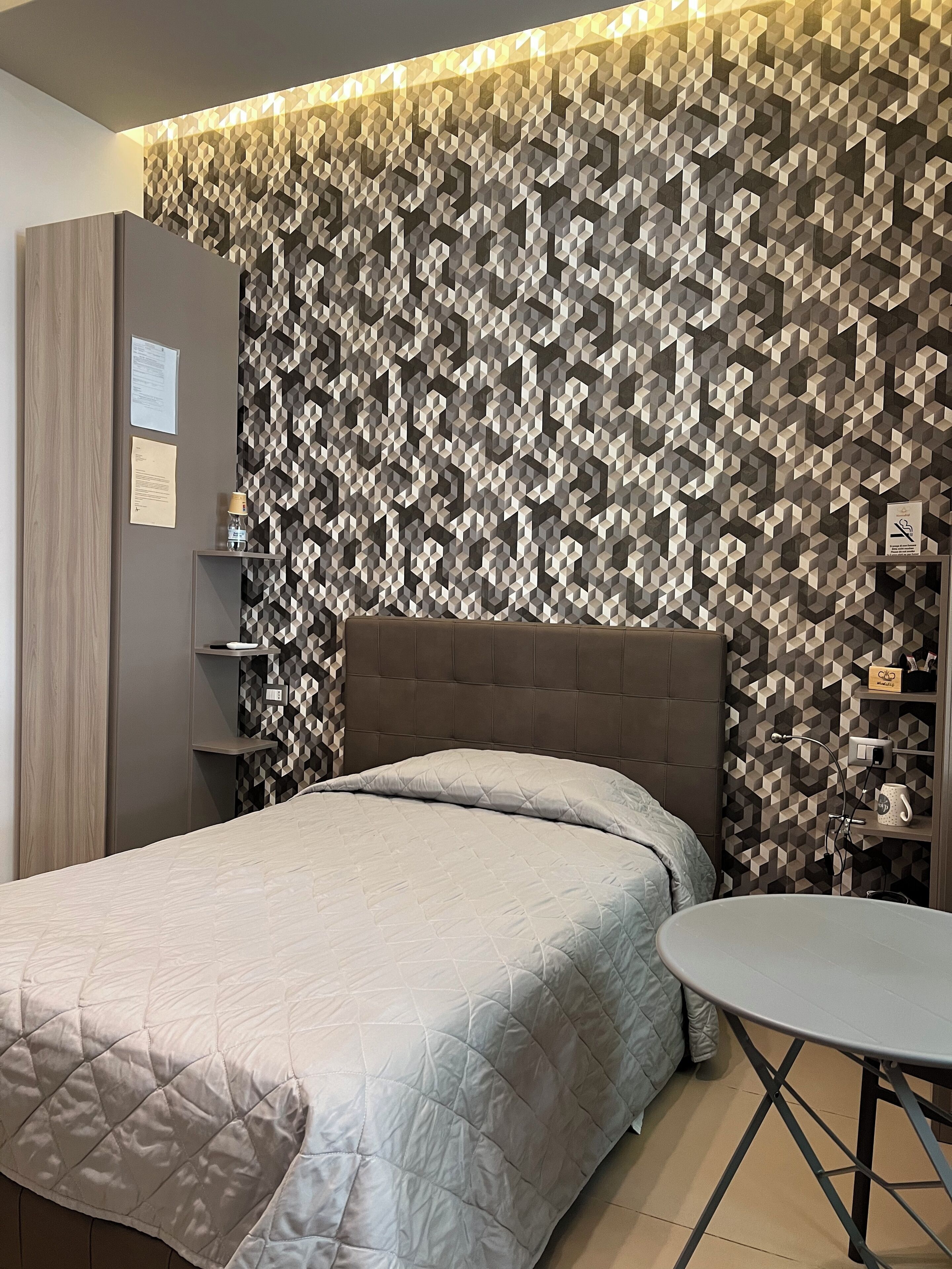 Un dormitorio moderno con pared de diseño geométrico, cabecera acolchada, cama con cubrecama acolchado, mesita de noche y una pequeña mesa redonda.