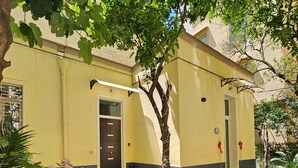 Un edificio amarillo con una puerta oscura, escalones que conducen a la entrada, y un árbol en el frente.