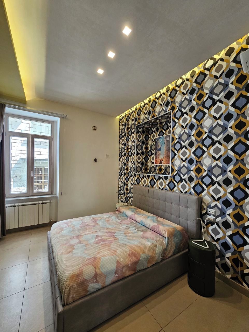A bedroom with a bed, a radiator, a window, and a patterned wall.