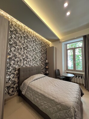 Quarto de hotel moderno com uma cama grande, uma mesinha e uma cadeira. Há uma janela com cortinas e vista para o verde lá fora.