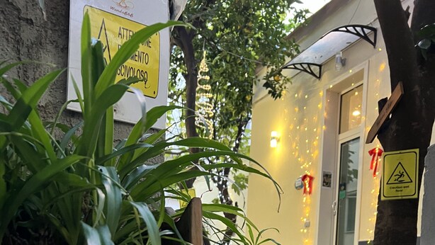 Una jardinera de madera con la leyenda 'Wanda B&B', un edificio amarillo con un letrero de piso mojado y una valla de madera con follaje.