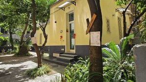 Un edificio amarillo con una puerta y escalones que conducen a él, rodeado de árboles y plantas.