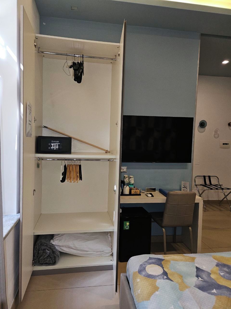 A compact room with a bed, a desk with a chair, a TV, and a wardrobe with hanging clothes.