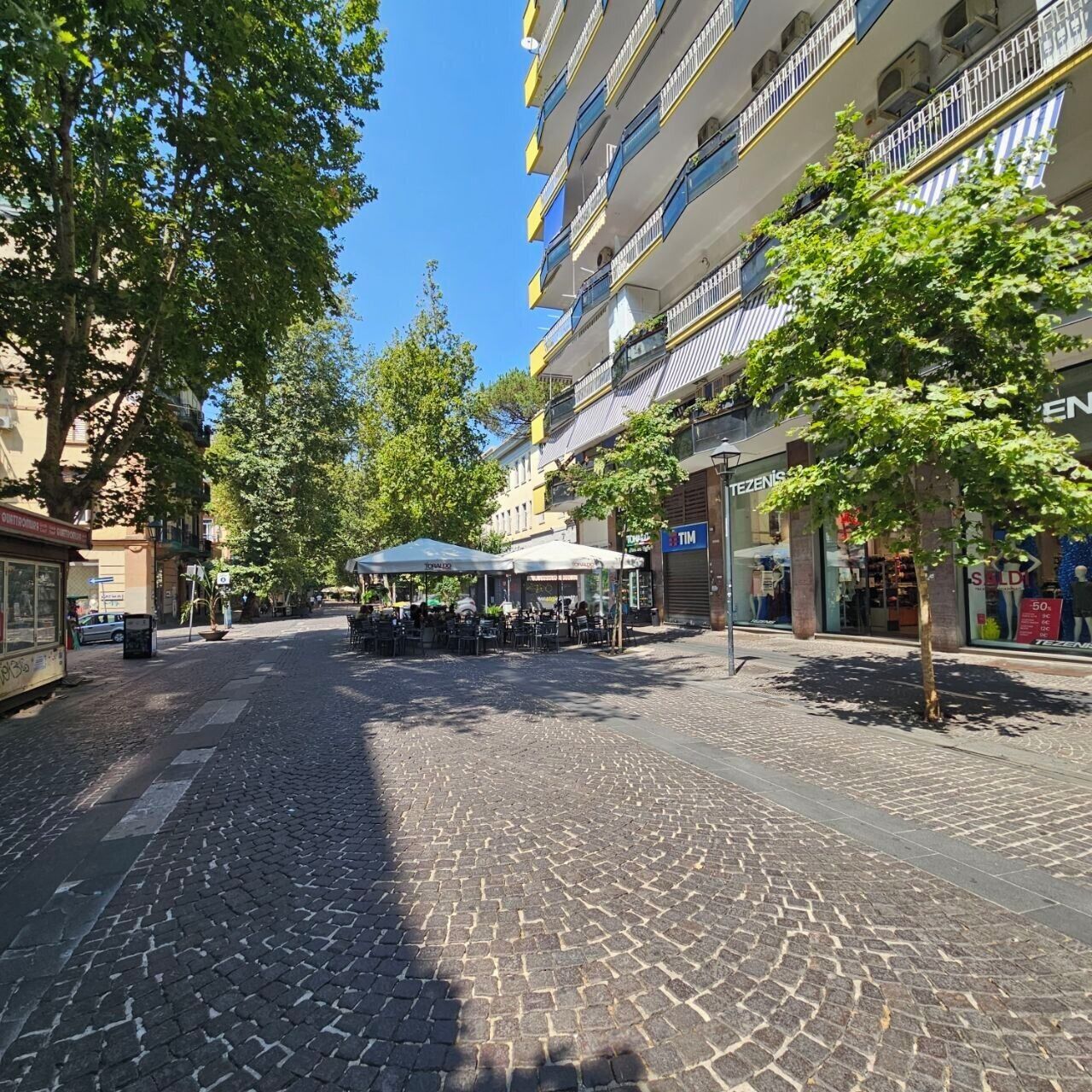 A cobblestone street with outdoor seating, storefronts, and a multi-story building with balconies.
