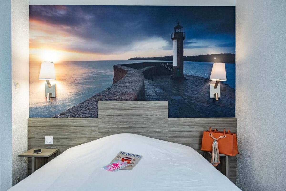 A hotel room with a large wall mural of a lighthouse, a bed with a magazine, and two bedside lamps.
