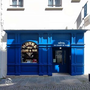 Eine Hotelansicht mit einem gut sichtbaren Schild 'HOTEL du CHAMP DE MARS', einer Kontaktnummer und traditioneller Architektur.
