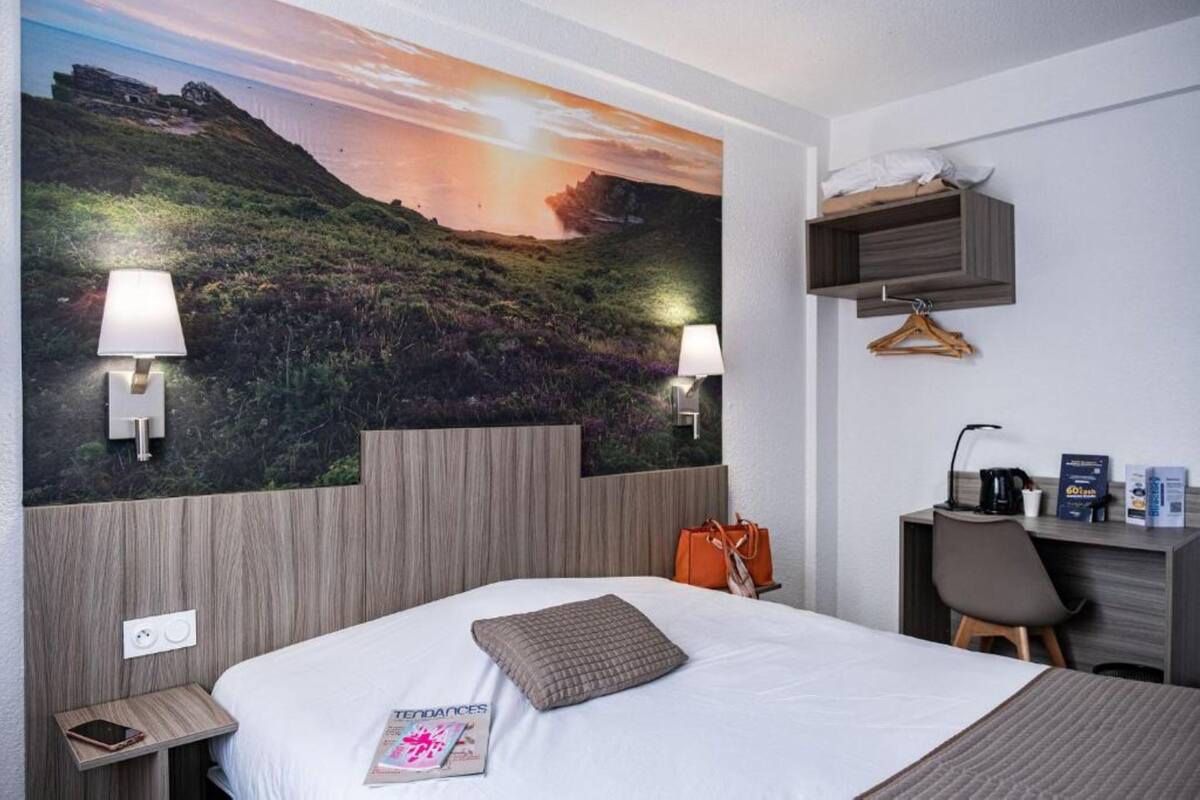 A hotel room with a large wall mural, a bed with a pillow, bedside lamps, a desk with a chair, and a hanging shelf.