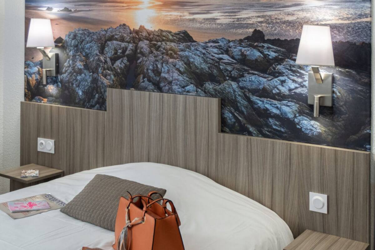 A hotel room with a large wall mural of a sunset over water, a bed with white linens, two bedside lamps, and a brown handbag on the bed.