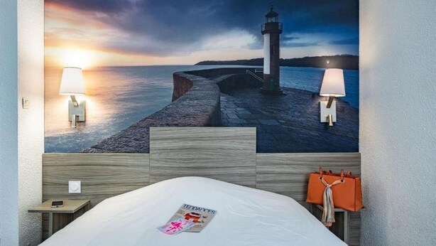 A hotel room with a large wall mural of a lighthouse, a bed with a magazine, and two bedside lamps. - Hôtel du Champ de Mars (Saint-Brieuc)