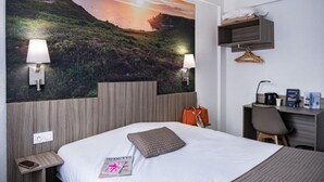 A hotel room with a large wall mural, a bed with a pillow, bedside lamps, a desk with a chair, and a hanging shelf. - Hôtel du Champ de Mars (Saint-Brieuc)