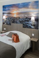 Habitación de hotel con un gran mural en la pared que muestra un atardecer sobre el agua, una cama con sábanas blancas, dos lámparas de noche y un bolso marrón sobre la cama.