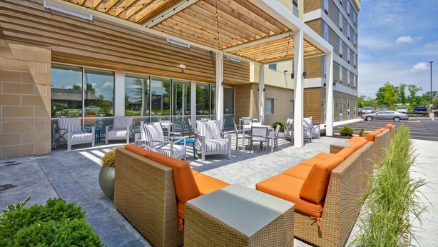 An outdoor seating area with wicker furniture and orange cushions, a covered patio with wooden beams, and a glass wall overlooking a parking area.