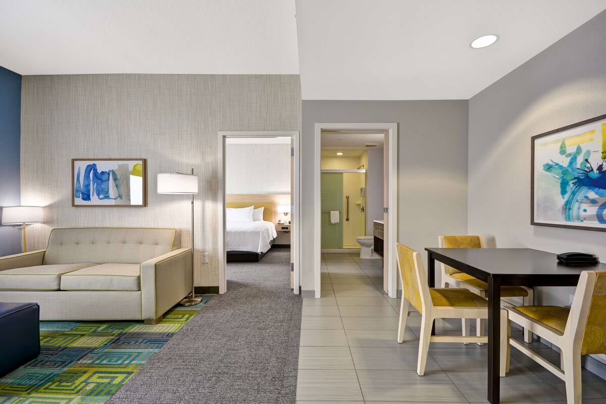A hotel room with a sofa, a dining table with chairs, a bed, and a bathroom visible through an open door.