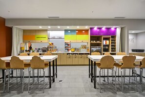A modern restaurant with a long communal table and individual chairs, a kitchen area with a colorful backsplash, and a counter with various food items and condiments.