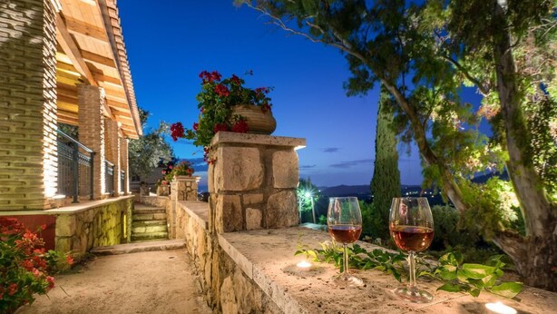 A stone pathway leading to a building with a stone pillar and a potted plant with red flowers. Two glasses of red wine are placed on the stone ledge. - Villa Aelia (Zakynthos)