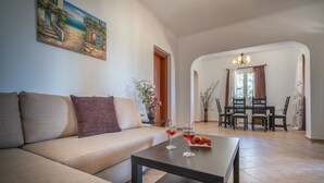 A living room with a sofa, a coffee table with wine glasses and strawberries, a painting on the wall, and a dining area with chairs and a chandelier. - Villa Aelia (Zakynthos)