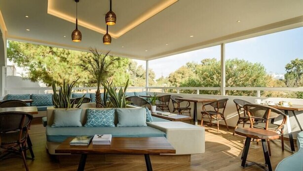 A modern outdoor seating area with a sofa, chairs, and tables, surrounded by large windows and plants. - Hotel Armonia (Santorini)