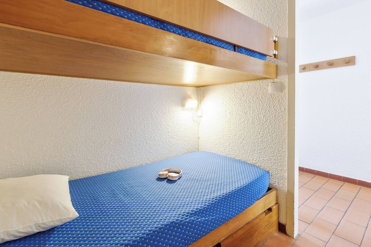 Un lit simple avec un matelas bleu et un oreiller blanc, une tête de lit en bois et un luminaire fixé au mur.
