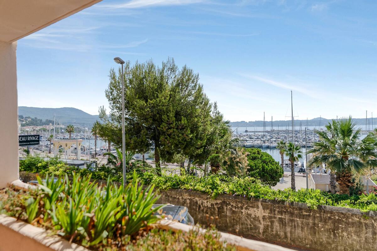 Une vue depuis un balcon donnant sur une marina, avec des bateaux, des palmiers et une chaîne de montagnes au loin.
