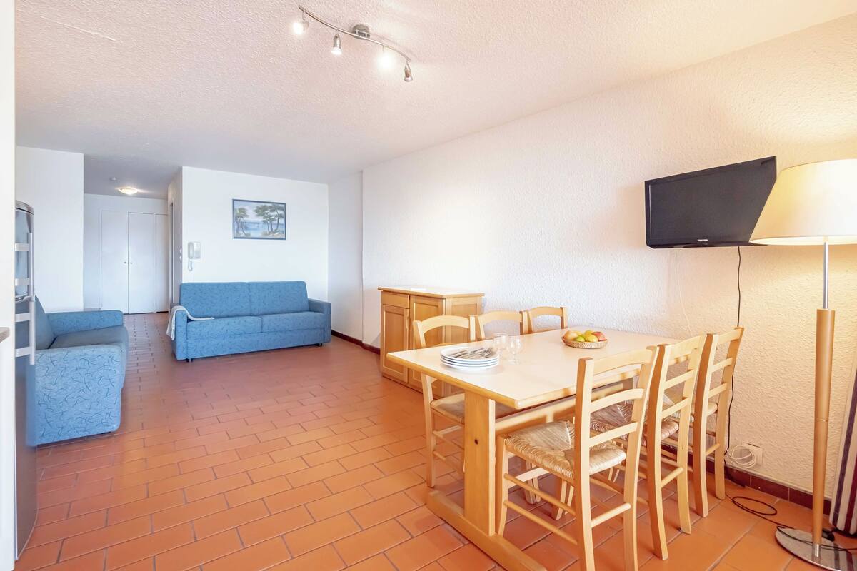 Un espace de vie compact comprenant un coin repas, un canapé, un téléviseur fixé au mur et un sol carrelé.