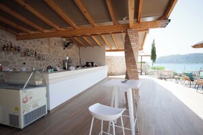 Zona de bar al aire libre con pared de piedra, vigas de madera y vista al mar.
