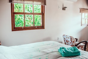 A room with a bed, a window with a view of greenery, a chair with a patterned cover, and a wall-mounted light switch. - Villa Seychelles Cabanas (Imbituba)