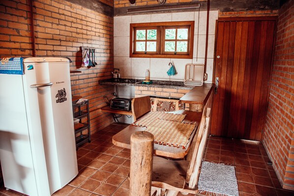 A compact kitchen with a refrigerator, wooden table, and chairs, a window with a view, and a brick wall. - Villa Seychelles Cabanas (Imbituba)