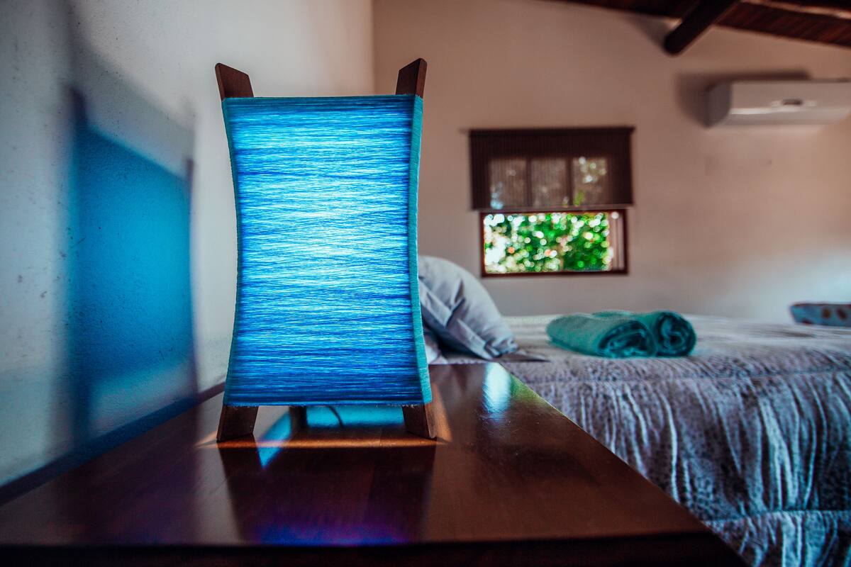 A bed with a blue blanket, a wooden nightstand with a blue object, and a window with greenery outside.