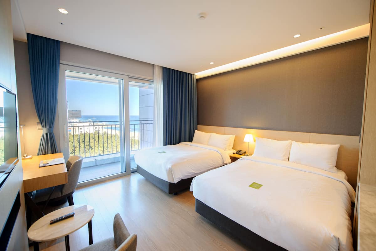 A hotel room with two beds, a desk, a chair, and a balcony with a view.
