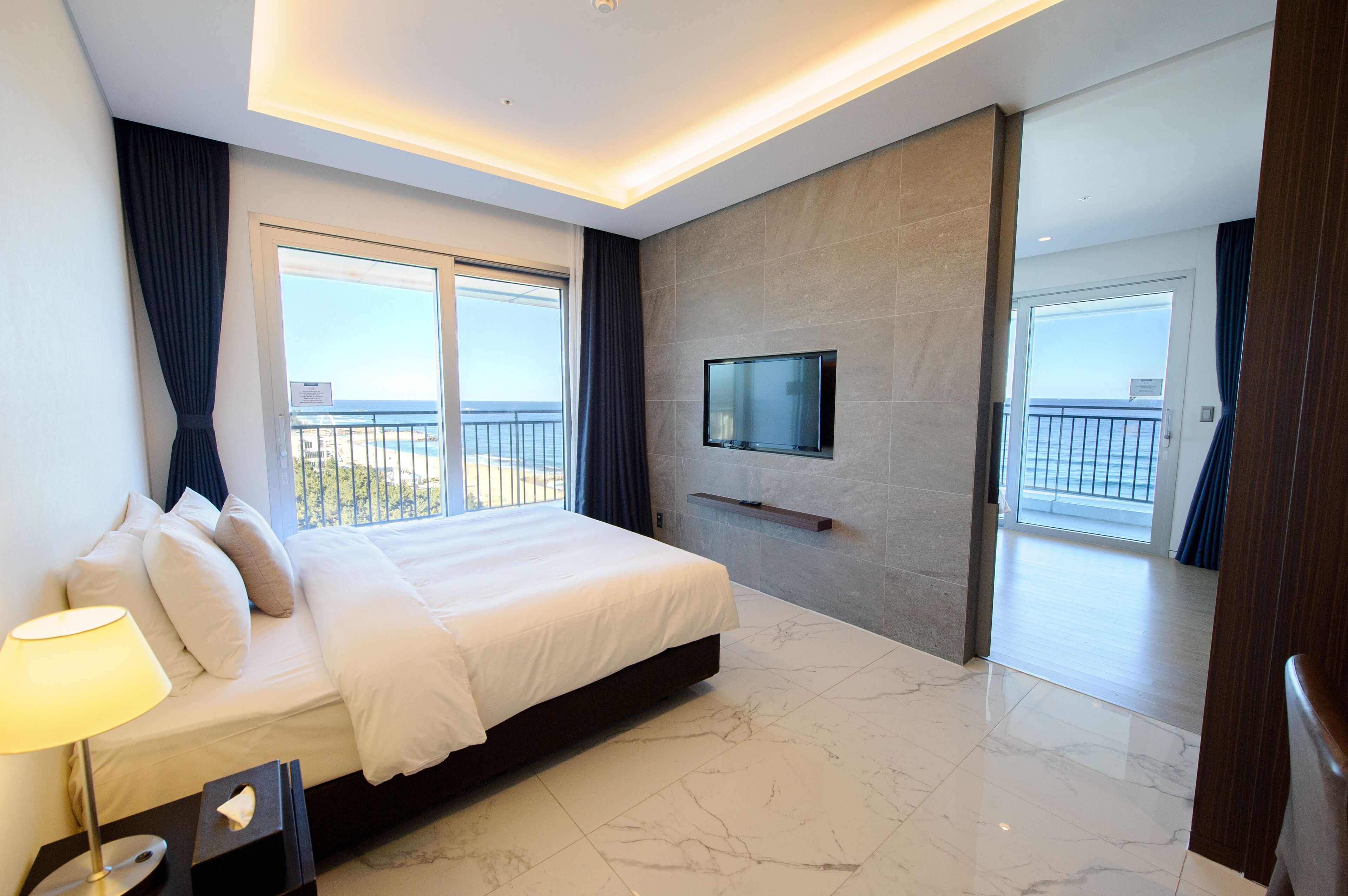 A modern hotel room with a large bed, a flat-screen TV mounted on the wall, and a balcony with a view of the sea.
