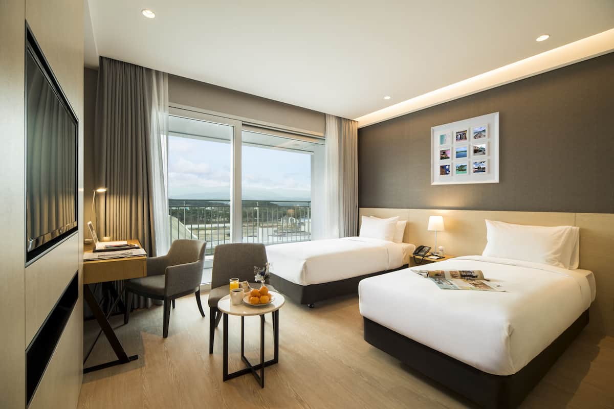 A hotel room with two beds, a desk with a laptop, a chair, a small table with a fruit bowl, and a window with a view of the city.