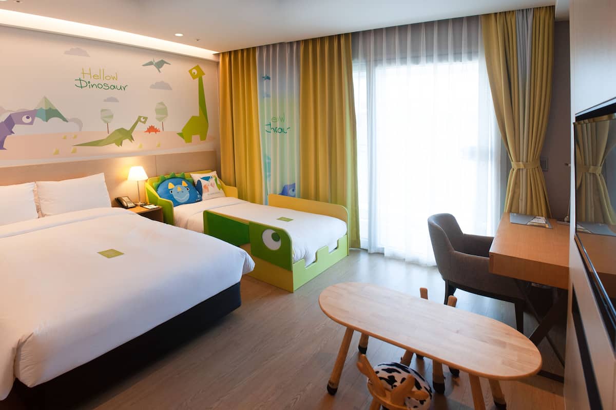 A hotel room with two beds, a desk, and a chair. There is a mural on the wall depicting dinosaurs and clouds.