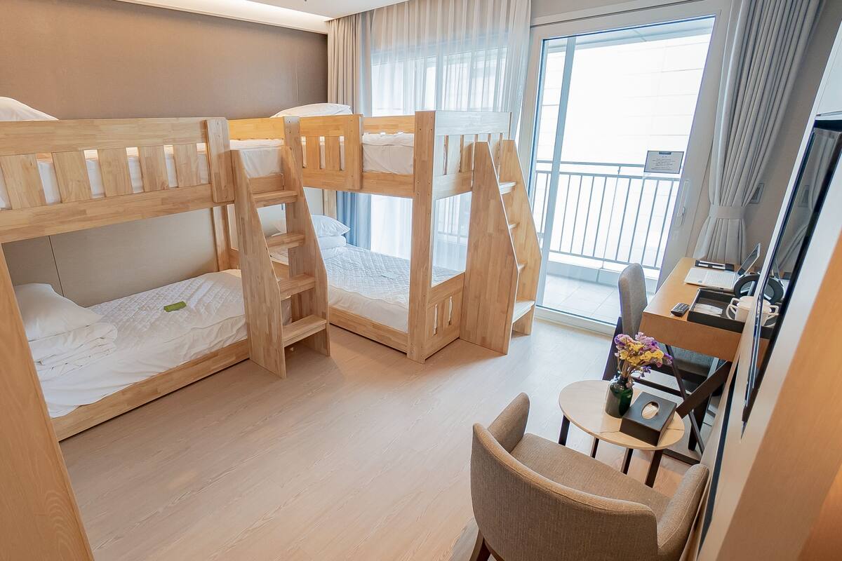 A bunk bed room with a desk and chair, a window with curtains, and a view of the outside.