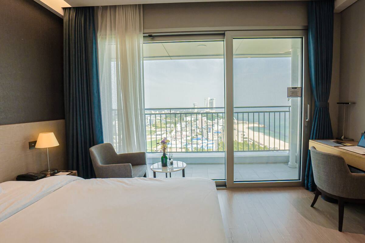A hotel room with a bed, a desk with a laptop, a chair, a small table with a vase, and a balcony with a view of buildings and a beach.